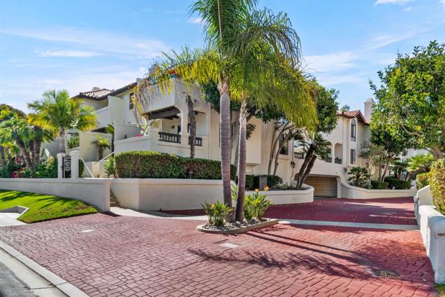 Entrance to Mediterranean-style townhome community with cream colored buildings, red cobblestone driveway, and palm trees.